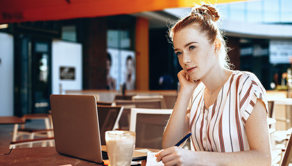 Red haired caucasian businesswoman with freckles writing and thinking about something in a cafeteria while drinking a cocktail and using a laptop