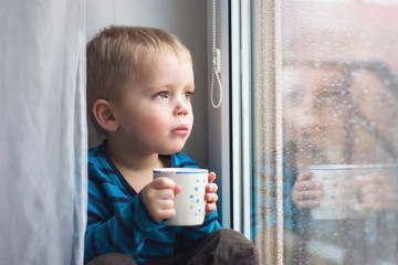 little boy with cup of tea sitting on the window and looking throgh the window in rainy day 