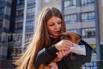 A girl trying to protect small dog from a coronavirus with a medical gauze white mask on the street in sunny spring day. Protection against disease during epidemics and pandemics. Covid-19 in Russia