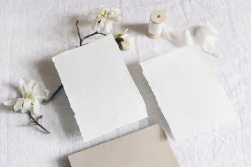 Obraz na płótnie Canvas Wedding stationery set. Mock-up scene with blank paper greeting cards, envelope on linen tablecloth background. White magnolia stellata tree branches and ribbon. Feminine still life, flat lay, top.