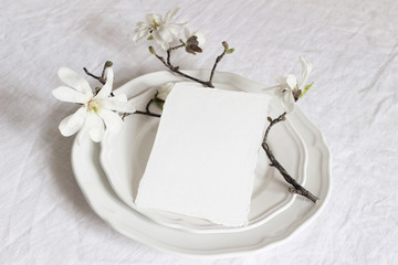 Festive table spring setting with porcelain plates, magnolia stellata tree branches with white blossoms and blank paper card mockup. Feminine composition, wedding or restaurant menu concept.