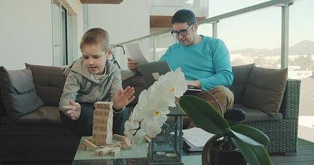 Stay and work at home dad. Father and son spend free time on terrace. Man working, child boy playing, builds, creating, wooden game toy. Weekend Family. Bonding concept. 4K video. - Powered by Adobe