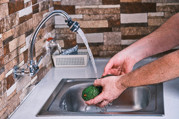 Man washing food due to coronavirus. Cleaning an avocado to remove viruses. Metalic sink and water to clean fruit.