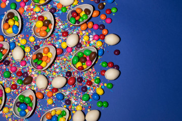 The left side of the photo is covered with halves of chocolate eggs, jellies and sweet powder. The right side of the photo is deep blue. Photo was taken by flat lay. There is a place for text.