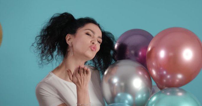 Portrait of beautiful young woman with black curly hair smiling and kissing towards the camera with ballons in her hands and blue background shot in 4k super slow motion