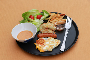 Breakfast in the black round plate on the brown floor. Fried egg with sausage, dumpling with french fries and vegetable with tomato.