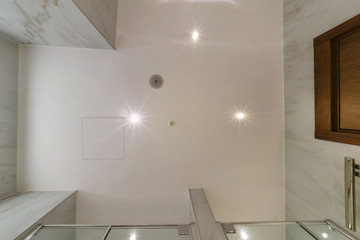 look up on suspended ceiling with halogen spots lamps and drywall construction with fire alarm sensor in empty room in apartment or house. Stretch ceiling white and complex shape.