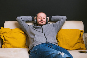 Bearded relaxed man in hoodie and trousers sitting on couch with headphones and listening music, smiling, eyes closed
