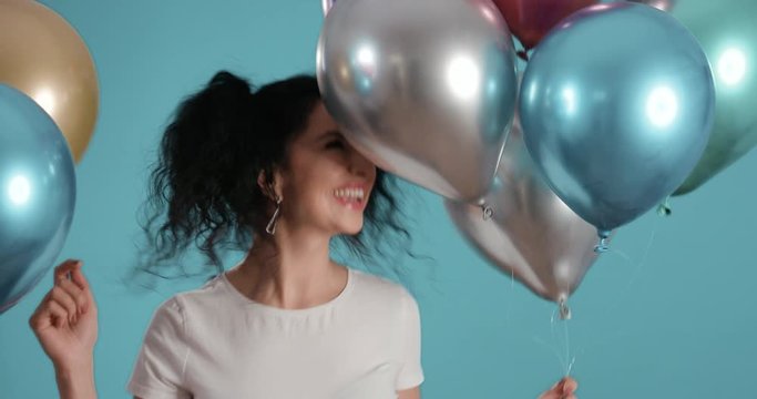 Portrait of beautiful young woman with black curly hair smiling and dancing with ballons in her hands in front of blue background shot in 4k super slow motion