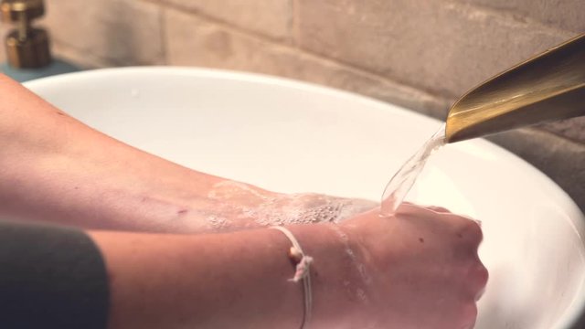 Young woman washing and rinsing hands with soap in a white oval sink bowl and brass fitted tap