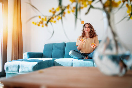Charming beautiful young girl with curly hair and a happy smile juggles tangerines while sitting on a blue sofa in her living room at home, having fun staying at home