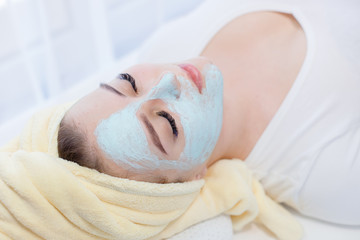 Fototapeta na wymiar Portrait of beautiful woman laying with towel on the head. Young girl enjoys cream facial mask. Lady getting spa treatment at beauty salon. Cosmetology and skincare concept.