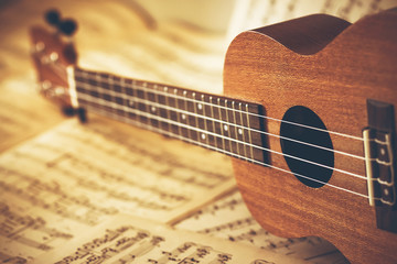 A ukulele with nylon strings lies on its side on scattered sheets of paper with notes on it. Music...