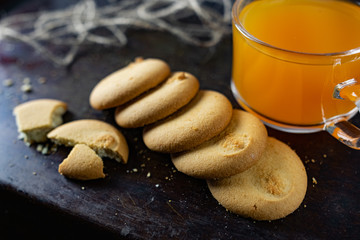 Homemade shortbread, round-shaped pastry on a dark old surface in a stack of crumbs. Next to it is a glass mug with orange mulfruit juice