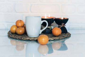 Fototapeta na wymiar Burning spa aroma candles in coconut shell, cup of tea, tangerines on wicker stand on a glass white table, cozy home interior background.