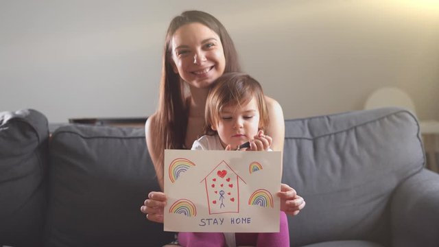 Happy young mother and baby on the sofa with drawing STAY HOME and rainbows. Global campaign for COVID 19
