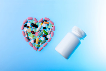 Multi-colored pills in the shape of heart and plastic jar on blue gradient background. Heart shape made of tablets, concept of treatment and healthcare. Copy space on the right side