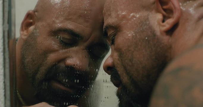 Bald, tan, muscular, bearded man pressing his head against a filthy mirror.