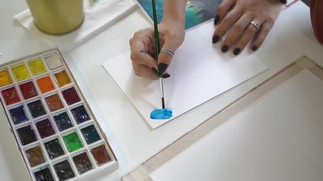 The girl draws on white paper with a brush and blue paint a drawing on the Mester class in the fine arts. Multi-colored watercolor palette with colored paints.