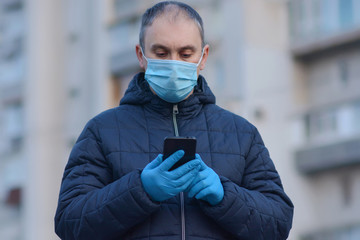Worried man in a protective sterile surgical mask and medical gloves checks his phone for news about the coronavirus outdoors
