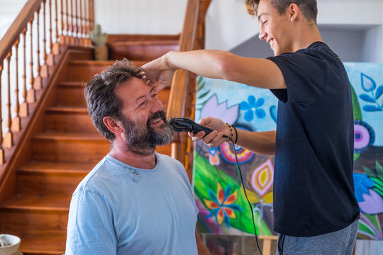 couple of dad and son at home laughing and having fun together cutting the beard of father in quarantine - smiling and playing at be a barber or hairdresser homemade
