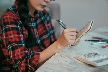 closeup view of woman's hand holding pen jotting down on notebook. girl sitting by table with workbook keeping track of spending with the help of calculator.