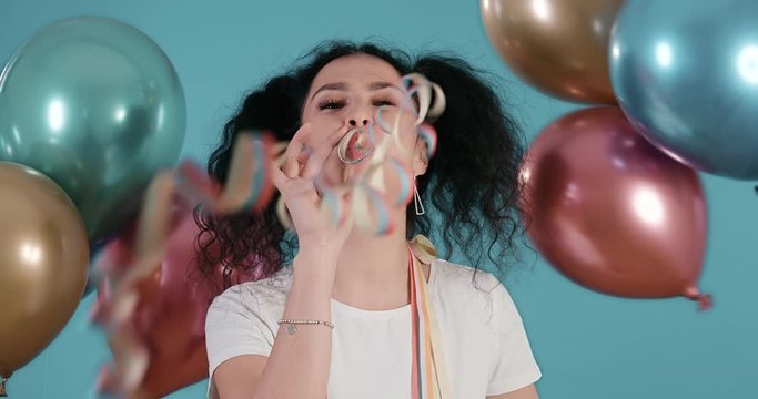 Portrait of beautiful young woman with black curly hair blowing streamers towards the camera with ballons in a hand and blue background shot in 4k super slow motion