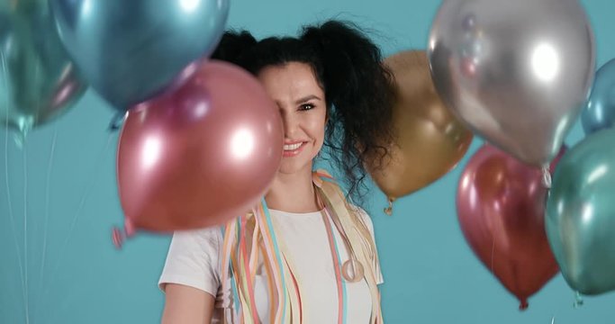 Portrait of beautiful young woman with black curly hair smiling and dancing with ballons in her hands in front of blue background shot in 4k super slow motion