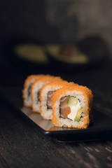 In a dark room on an old table stands sushi with orange caviar shrimp and salmon on a black plate, in the background you can see a whole and sliced avocado