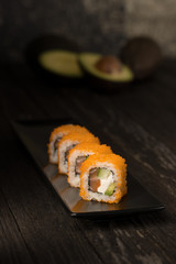 In a dark room on an old table stands sushi with orange caviar shrimp and salmon on a black plate, in the background you can see a whole and sliced avocado
