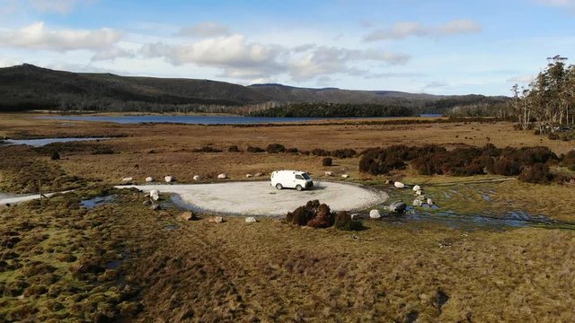 Fast drone shot downwards into campervan in wilderness surrounded by bushland, lakes and mountains