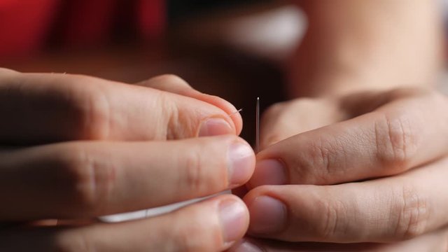 Threading the eye of a needle. Pulling the thread into the needle eye of the. Young man pulling the white thread into the needle eye of the sewing needle at night in home. Close up.