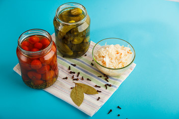 Jars with pickled cucumbers and tomatoes and sauerkraut on a wooden table on a blue background on a linen towel.