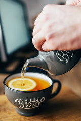 Fototapeta na wymiar A man pours tea from a ceramic teapot into a ceramic cup - a tea break when working in the office at the computer table