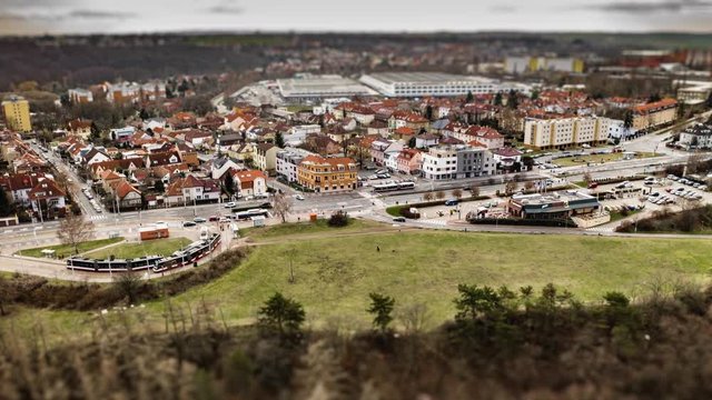 Aerial view of a tram stop and a crossroad near Divoka Sarka park in Prague. Active traffic on the road, cars, busses, and trams. Commuters crowd the tram stop. People walking dogs in the park nearby