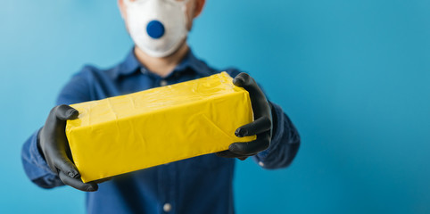 A young delivery man in a medical mask holds a parcel box in his hands on a blue background. Banner. The concept of courier delivery.