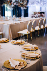 Emty glasses for wine and water are on the table, there are golden plates with napkins, the table is covere with beige tablecloth, guests chairs on the background