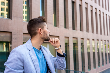 Pensive bearded young man leaning on a fence with hand on chin while looking away