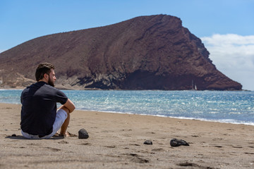 A young handsome man sitting on the sand in a beach while looking at the sea, in Tenerife, Canary Islands, Spain