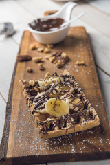 Waffle with banana slices, oatmeal, nuts, and chocolate served on a wooden cutting board over a white rustic wooden table. Hazelnut chocolate spread in the background. Closeup.