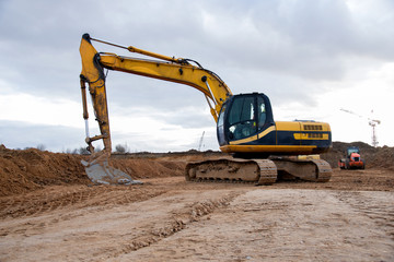 Yellow excavator during earthworks at construction site. Backhoe digging the ground for the foundation and for laying sewer pipes district heating. Earth-moving heavy equipment