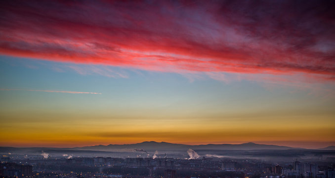 Fotografía de Burgos al amanecer, con las montañas de la Sierra de la Demanda al fondo y bajo las nubes de color rojo. Junto a las montañas se distingue el humo de las fábricas