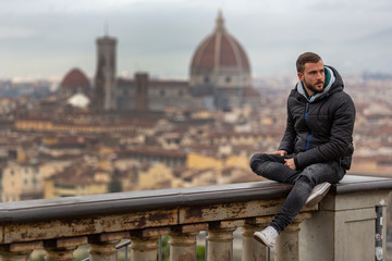 A young handsome man with beard sitting on a wall in a viewpoint of Florence, Italy