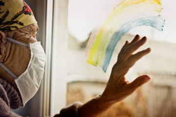 An old grandmother in a protective mask looks out the window at self-isolation. Elderly woman at quarantine at home. Grandmother looks through the rainbow on the window pane into the street with a clo
