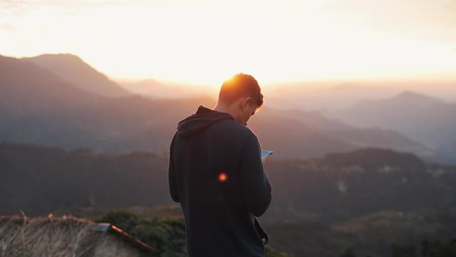 A man looking at his phone while having a beautiful mountain landscape view at sunset, sunrise.