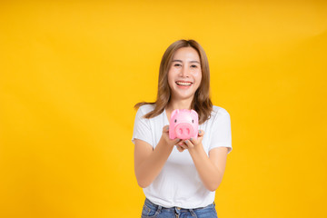 Portrait happy young asian woman holding piggybank saving money for future and business earning income positive emotion in white t-shirt, Yellow background isolated studio shot and copy space.