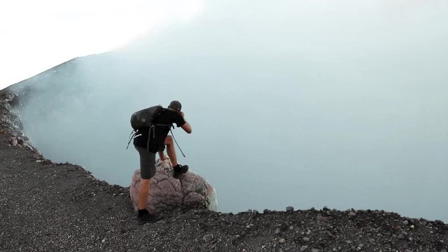 Wide shot of a middle aged white caucasian man wearing a backpack standing at the edge of the active Telica volcano looking inside the sulfur smoke filled crater while resting on foot on a big rock