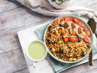 Delicious amaranth porridge topped with home made granola, strawberries and almond butter. Vegan plant based breakfast served with matcha latte on white wooden board. Healthy colorful food concept