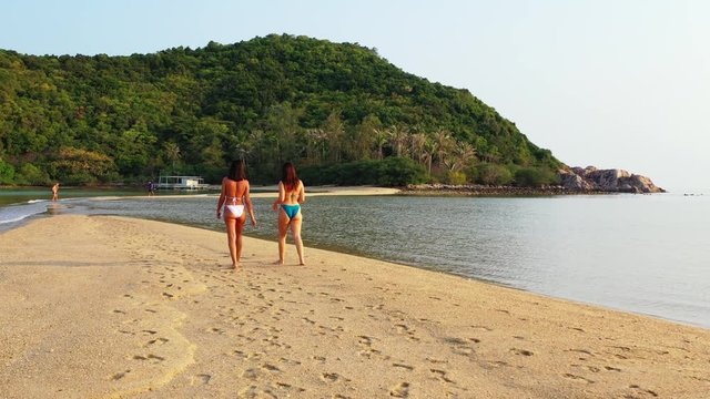 Following shot of young women in swimsuit walking on sandy beach washed by calm lagoon reflecting sunlight on tropical island in Vietnam