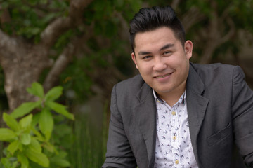Happy young overweight Asian businessman smiling at the park